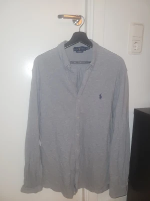 Grå skjorta från Ralph Lauren - Säljer en klassisk grå skjorta från Ralph Lauren med blå logobrodyr på bröstet. Skjortan har lång ärm, knappar framtill och är tillverkad i mjuk bomull. Perfekt för dig som gillar stilrena plagg med ikonisk design.