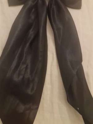 Stor svart rosett hårklämma - Snygg och stor svart rosett i glansigt tyg med metallklämma på baksidan. Perfekt för att ge en extra touch till din frisyr. Rosetten har långa band och är gjord i ett tunt, elegant material som ger ett lyxigt intryck.