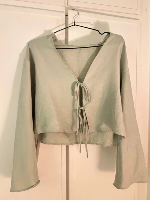 Ljusgrön croppad topp med knyt - Trendig ljusgrön croppad topp med långa ärmar och två knyt framtill. Toppen har v-ringning och är tillverkad i ett mjukt, tunt tyg som ger en avslappnad look. Perfekt att styla med höga jeans eller kjol.