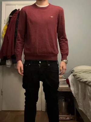 Bordeaux stickad tröja från GANT - Säljer en stilren stickad tröja från GANT i färgen Bordeaux Melange. Tröjan har rund halsringning, ribbade muddar och diskret logga broderad på bröstet. Perfekt för dig som gillar klassisk och enkel stil.