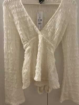 Vit omlottopp med volang och knyt - Supersöt vit omlottopp från Monki i smockat tyg med långa ärmar och volangkant. Toppen har v-ringning och knyt i midjan för en snygg passform. Perfekt att styla med jeans eller kjol för en trendig look.