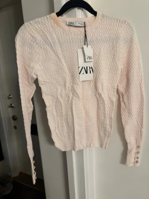 Ljusrosa stickad tröja från ZARA - Trendig ljusrosa stickad tröja från ZARA med strukturerat mönster och rund hals. Långa ärmar med dekorativa knappar vid ärmsluten ger en extra detalj. Perfekt för dig som gillar stilrena och feminina plagg. Materialet känns mjukt och bekvämt.
