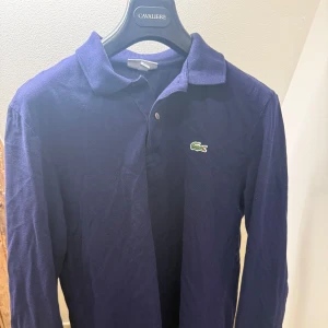 Mörkblå långärmad pike Lacoste - Klassisk mörkblå långärmad skjorta från Lacoste med ikoniska krokodilloggan på bröstet. Skjortan har krage, knappar framtill och är tillverkad i ett mjukt bomullsmaterial. Perfekt för en stilren och avslappnad look. Storlek M.