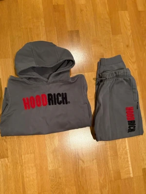 Grå HOODRICH hoodie + byxor - Säljer en grå hoodie från HOODRICH med stor broderad logga i rött och svart på bröstet. Matchande gråa byxor med samma logga på benet.