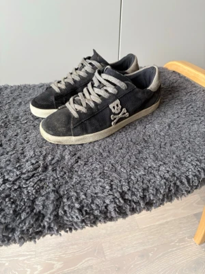 Svarta sneakers med dödskalle - Svarta låga sneakers med gråa skosnören och vit sula. Sidan pryds av en broderad dödskalle och korslagda ben. Skorna har klassisk siluett och är tillverkade i canvasmaterial. Perfekta för dig som gillar en edgy och avslappnad stil.