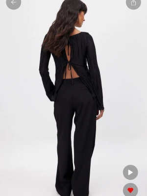 Svart plisserad topp med öppen rygg - Trendig svart plisserad topp med långa ärmar och öppen rygg med snygga knytband. Toppen har en rak passform och är tillverkad i ett lätt, glansigt material som ger en lyxig känsla. Perfekt för dig som vill sticka ut med en cool detalj i ryggen. Aldrig använd med prislappen kvar - originalpris: 399 kr