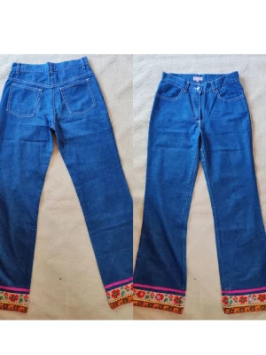 Blå jeans med broderad kant - Unika blå jeans med raka ben och färgglad broderad kant längst ner. Dekorerade med blommor, mönster i rött, grönt, gult och rosa samt en rosa banddetalj. Perfekta för dig som vill sticka ut med en bohemisk och kreativ stil.