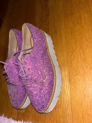 Glittriga lila platåloafers - Säljer ett par loafers med lila glitter och tjock vit platåsula. Skorna har snörning framtill och en cool, ungdomlig vibe. Perfekta för dig som vill sticka ut med färg och glitter. Materialet är syntet med glittrig yta och innersula i beige textil.