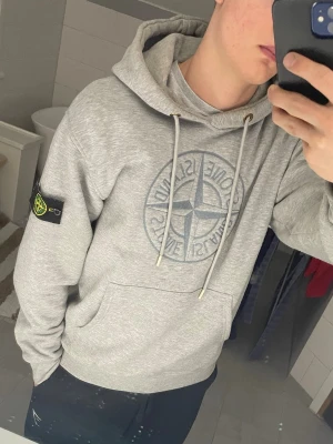 Grå hoodie från Stone Island - Säljer en grå hoodie från Stone Island med stor logga på bröstet och den klassiska patchen på vänster ärm. Hoodien har huva med dragsko och en stor magficka. Tillverkad i mjukt bomullsmaterial och har ribbade muddar vid ärmslut och nederkant.