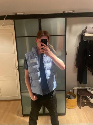 Hoodrich väst  - Snygg ljusblå dunväst från Hoodrich med dragkedja framtill och två fickor. Västen har en hög krage och tydlig logga på bröstet. Perfekt för lager-på-lager och streetwear-stil. fins en lite fläk men den syns knappt 