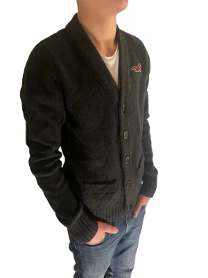 Mörkblå kofta från Hollister - Snygg svart kofta från Hollister med knappar framtill och två fickor. Koftan har ribbade muddar och ett broderat Hollister-logo på bröstet. Perfekt att slänga på över en t-shirt för en chill look. Modellen är 178cm,  9/10 skick