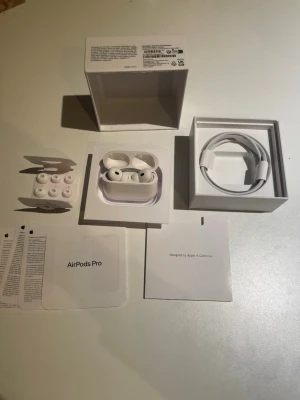 Apple AirPods Pro trådlösa hörlurar - Säljer ett par vita Apple AirPods Pro med laddningsetui, extra silikonpluggar och laddningskabel. Hörlurarna har en stilren och kompakt design i plast och levereras med originalförpackning och manualer. Perfekta för dig som vill ha grymt ljud och smidig anslutning.
