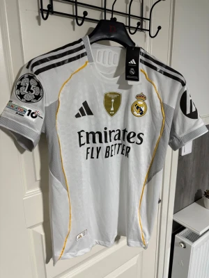 Real Madrid matchtröja 2024 Adidas - Snygg vit Real Madrid matchtröja från Adidas med svarta och gula detaljer. Tröjan har klubbmärke, FIFA-märke och Champions League-märke på ärmen. Tillverkad i lätt och ventilerande material, perfekt för fotboll eller träning.