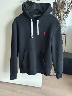Svart hoodie från Polo Ralph Lauren - Svart hoodie från Polo Ralph Lauren med vit dragsko i huvan och röd broderad logga på bröstet. Klassisk känguruficka framtill och ribbade muddar vid ärmslut och nederkant. Perfekt för en avslappnad och stilren look.