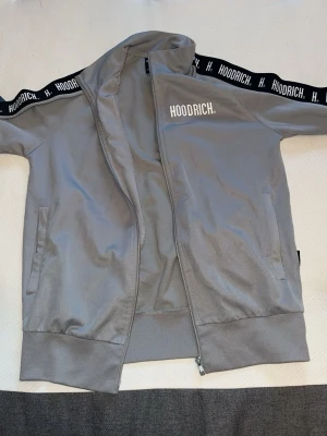 Grå hoodie från Hoodrich med dragkedja - Snygg grå hoodie från Hoodrich med dragkedja och huva. Svarta band med Hoodrich-logga längs axlarna och ärmarna. Två fickor framtill och ribbade muddar. Perfekt för en streetwear-look.