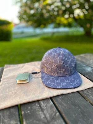 Blå keps från Gbg med GG-mönster - Säljer en snygg lila keps från Gg med det klassiska GG-monogrammet över hela kepsen. Kepsen har böjd skärm och justerbar rem bak. Materialet är canvas och kepsen kommer med originalpåse och taggar kvar.