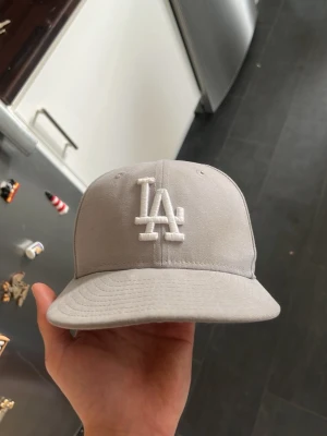 Grå LA-keps från New Era - Säljer en stilren grå keps från New Era med LA-logga broderad i vitt framtill. Klassisk modell med böjd skärm och broderade ventilationshål. Perfekt accessoar för dig som gillar streetwear och sportig stil.