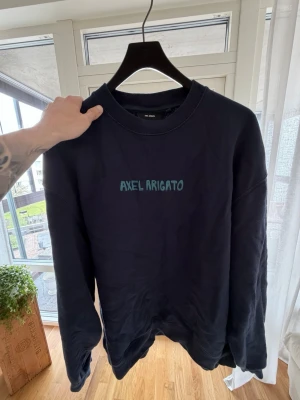 Mörkblå sweatshirt Axel Arigato - Säljer en mörkblå sweatshirt från Axel Arigato med rund halsringning och broderad logga i turkost framtill. Tröjan har en avslappnad passform och ribbade muddar vid ärmslut och nederkant. Perfekt för en casual och stilren look.