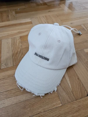 Vit keps från Jacquemus - Stilren vit keps från Jacquemus med metallogo framtill. Kepsen har böjd skärm med slitna kanter och justerbar rem baktill. Perfekt accessoar för en trendig och avslappnad look.