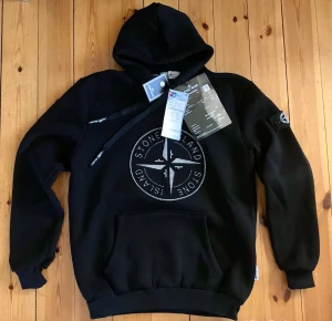 Svart hoodie från Stone Island - Svart hoodie från Stone Island med stor broderad logga på bröstet och klassisk patch på vänster ärm. Huva med dragsko och magficka framtill. Perfekt för dig som gillar streetwear och vill ha något stilrent men ändå med attityd.