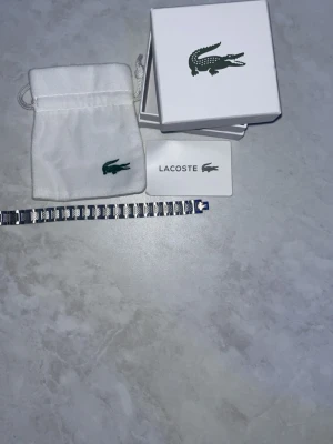 Silverfärgat armband från Lacoste - Snyggt armband i silverfärgad metall från Lacoste med rektangulära länkar och den klassiska krokodil-loggan. Armbandet har en stilren och modern design och levereras med originalask och tygpåse.