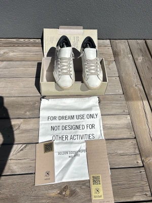 Vita Golden Goose sneakers i läder - Stilrena vita sneakers från Golden Goose med klassisk låg siluett. Skorna är tillverkade i läder med gråa skosnören och har diskreta detaljer som stjärna på sidan och svart hälparti. Perfekta för en trendig och avslappnad look. Box och allt annat får man med sig i köpet. 