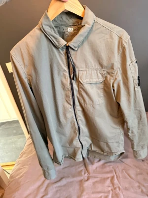 Mörkgrön overshirt från C.P. Company - Snygg mörkgrön overshirt från C.P. Company med dragkedja framtill, bröstficka och klassisk krage. Långärmad modell i bomull med en ikonisk detalj på ärmen. Perfekt lager-på-lager-plagg för en clean och stilren look. Köpt på CareOfCarl för ca 3 år sedan. Säljer då jag växt ur den. 