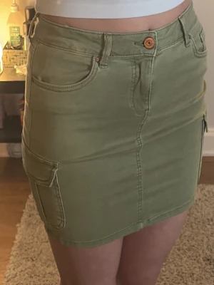Grön kort cargo-kjol med fickor - Cool grön cargo-kjol med stora sidofickor och klassiska jeansdetaljer som knapp och hällor. Kjolen är kort och har en rak passform, perfekt för dig som gillar en avslappnad och trendig stil. Materialet är bomull och ger en skön känsla.