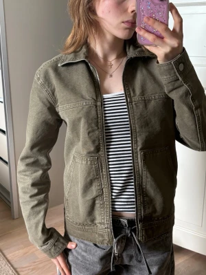 Olivgrön jeansjacka från Monki - Olivgrön jeansjacka från Monki med två stora fickor framtill. Jackan har klassisk krage, dragkedja och är tillverkad i kraftigt jeanstyg. Perfekt att slänga över en t-shirt för en avslappnad look.