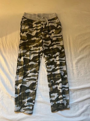 Camo mjukisbyxor från KappAhl för barn  - Säljer ett par mjukisbyxor från KappAhl med grön, grå och vit camomönster. Byxorna har resår och dragsko i midjan samt muddar vid bensluten. Perfekta för chill dagar och riktigt sköna att ha på sig.