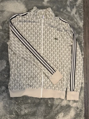 Mönstrad zip hoodie från Lacoste Storlek M - Säljer en snygg beige och vit zip hoodie från Lacoste med diskret logga på bröstet. Hoodien har ett rutigt mönster, ribbade muddar och svarta samt vita ränder längs ärmarna. Tillverkad i mjukt material som känns skönt mot huden. Perfekt för en sportig och trendig look. Storlek M
