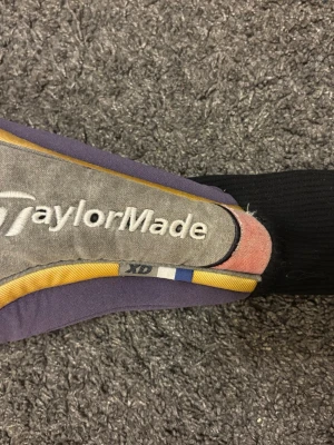 TaylorMade golfklubbskydd - Säljer ett TaylorMade skydd för golfklubba i tyg med broderad logga. Skyddet är mestadels grått och lila med detaljer i gult, blått och rosa. Passar perfekt för att skydda klubbhuvudet i bagen.