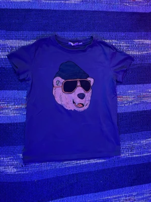 Blå t-shirt med tryck - En blå t-shirt med korta ärmar och ett stort grafiskt tryck framtill som föreställer ett tecknat björn ansikte med mössa. T-shirten har en klassisk passform och rund halsringning. Perfekt för dig som gillar streetwear och coola prints.