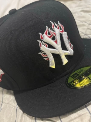 New Era Yankees keps svart 59FIFTY - Svart New Era 59FIFTY keps med New York Yankees-logga i vitt med röda flames framtill och broderad World Series 1996-patch på sidan. Klassisk flat peak och broderade detaljer. Perfekt för dig som vill sticka ut med streetstyle.
