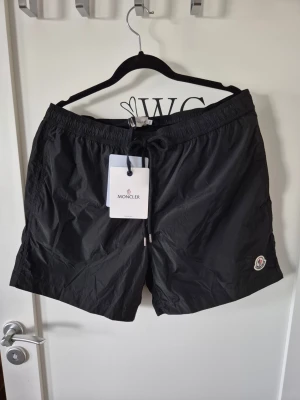 Svarta shorts från Moncler med snörning L - Svarta Moncler shorts med elastisk midja och snörning framtill. Klassisk Moncler-logga på vänster ben. Lätta och snygga shorts i syntetmaterial, perfekta för sommaren. Passar en i storlek med W32/33