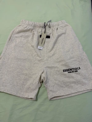 Essentials Fear of God shorts grå - Snygga grå shorts från Essentials Fear of God med elastisk midja och logga tryckt på ena benet. Perfekta för en avslappnad stil och sköna att bära tack vare mjukt bomullsmaterial.
