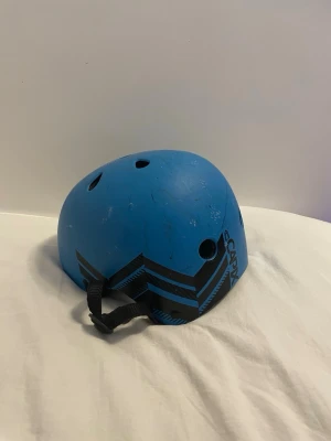 Blå skatehjälm SCAMPX - Säljer en blå skatehjälm från SCAMPX med svarta grafiska detaljer på sidan. Hjälmen har flera ventilationshål och justerbart hakband. Perfekt för skate, cykel eller kickbike. Tillverkad i tålig plast med bekväm passform.