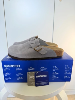 Grå Birkenstock Boston tofflor - Säljer ett par snygga Birkenstock Boston tofflor i mörkgrå mocka med justerbart spänne och klassisk korksula. Perfekta för dig som vill ha både stil och komfort hemma eller ute. Modellen har en mjuk innersula och tidlös design.