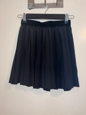 Svart plisserad kjol från H&M Divided - Snygg svart plisserad kortkjol från H&M Divided. Kjolen har en elastisk midja och är tillverkad i ett lätt och luftigt material som ger ett fint fall. Perfekt att styla med en t-shirt eller stickad tröja.