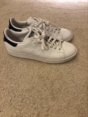 Adidas Stan Smith vita sneakers storlek 43 - Klassiska vita Adidas Stan Smith sneakers med svarta detaljer på hälen. Skorna har en låg siluett, perforerade ränder på sidorna och är tillverkade i slätt läder med vit gummisula. Perfekta för en clean och stilren look. Änvend Max 3 gånger ny pris 1200 kr mitt pris 500