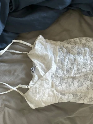 Vit spetslinne med smala axelband - Säljer ett vitt linne från new yorkermed smala axelband och v-ringning. Linnet är täckt av vacker spets och har en feminin och fräsch look. Perfekt att styla med jeans eller kjol för en trendig outfit.