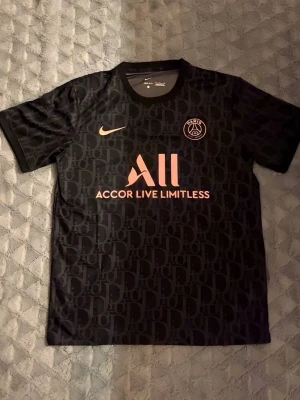 Svart PSG x Nike matchtröja - Snygg svart PSG matchtröja från Nike med diskret mönster, rosa logga och texten 'Accor Live Limitless' på bröstet. Tröjan har rund hals och korta ärmar, perfekt för fotboll eller streetwear. Tillverkad i lätt och ventilerande material.