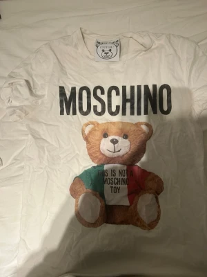 Vit Moschino t-shirt med björntryck - Vit t-shirt från Moschino med stort tryck av en nallebjörn och texten 'THIS IS NOT A MOSCHINO TOY' på framsidan. T-shirten har rund hals och korta ärmar. Perfekt statement-plagg för dig som gillar lekfull design och streetstyle.