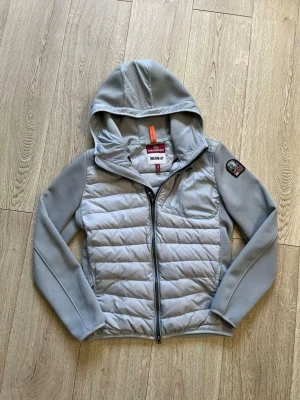 Ljusgrå pufferjacka med huva - Snygg ljusgrå pufferjacka från Peak Performance med quiltad front och rygg, samt släta ärmar och huva. Jackan har dragkedja framtill, en bröstficka och en patch på ena ärmen. Perfekt för kalla vinterdagar.