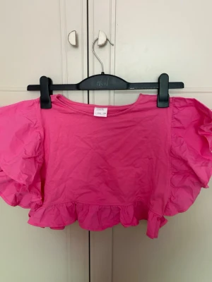 Rosa volang topp - Säljer en söt volang topp från Zara i stark rosa färg. Toppen har korta, voluminösa ärmar och volangdetalj längs nederkanten. Perfekt för dig som vill sticka ut med en färgglad och trendig look.