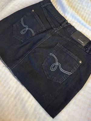 Svart jeanskjol från October - Snygg svart jeanskjol från October med klassisk femficksdesign och dekorativa vita sömmar på bakfickorna. Kjolen har en rak, kort passform. Midjemått: 38 cm rakt över (lite stretchig)