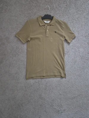 Olivgrön piké - Snygg beige pikétröja från H&M med klassisk krage, kort ärm och knappslå framtill. Liten broderad logga på bröstet. Perfekt till jeans eller shorts för en clean look. 