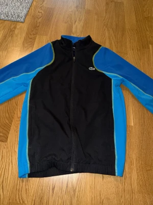 Svart och blå träningsjacka Lacoste - Sportig träningsjacka från Lacoste Sport i svart med blå ärmar och gröna detaljer. Jackan har hel dragkedja framtill, ståkrage och det klassiska Lacoste-krokodilmärket på bröstet. Perfekt för träning eller som en snygg streetwear-detalj.