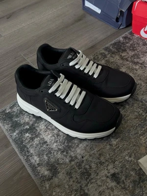 Svarta Prada sneakers med vit sula - Snygga svarta sneakers från Prada med vit tjock sula och vita skosnören. Skorna har en klassisk låg siluett och ikonisk Prada-logga på sidan. Tillverkade i en mix av textil och läder för en exklusiv känsla och modern look.