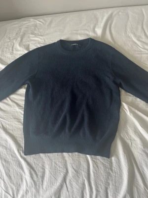 Mörkblå stickad tröja från J.Lindeberg - Snygg mörkblå stickad tröja från J.Lindeberg med rund halsringning och ribbade muddar. Tröjan har en klassisk, rak passform och är tillverkad i ett mjukt, strukturerat material som passar perfekt till både jeans och chinos.